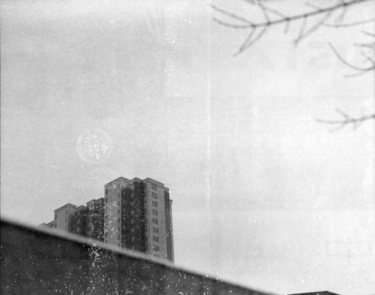 Xiamen Pan 100 (6x7)

English

A black-and-white photograph showing a cityscape during snowfall. The foreground features a rooftop with a thin layer of snow, and in the background, there are tall apartment buildings partially obscured by the snow. The sky is overcast, and snowflakes are visible in the air.

中文

一张黑白照片，拍摄的是下雪天的城市景观。前景是一个屋顶，上面覆盖着一层薄雪，背景是几栋高层公寓楼，部分被雪花遮挡。天空阴沉，空中可见飘落的雪花。