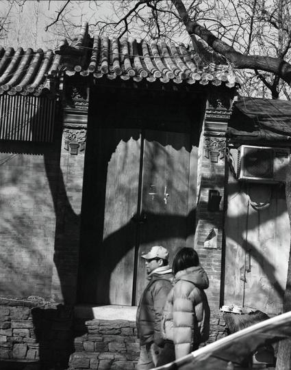 Lucky LUCKY SHD 400 (6x7)

English

A black-and-white photograph showing two people walking past a traditional Chinese building entrance. The entrance has a tiled roof and a large wooden door. The walls are constructed from gray bricks, and an air conditioning unit is mounted on the right side. Shadows of tree branches are visible on the door and the ground.

中文

一张黑白照片，照片中有两个人走过中国传统建筑的入口。入口处有瓦屋顶和一扇大木门。墙是用灰砖砌成的，右侧墙上安装有一台空调。树枝的影子投射在门和地面上。
