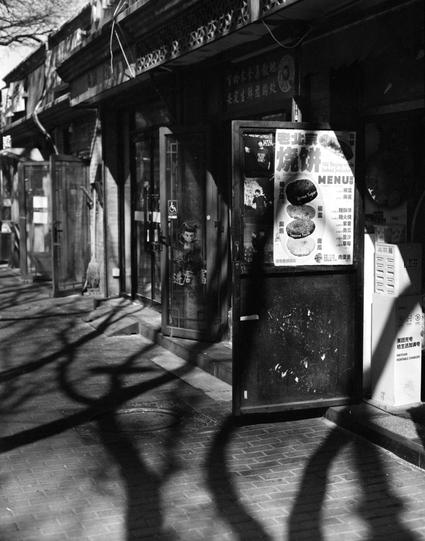 Lucky LUCKY SHD 400 (6x7)

English

A black-and-white photograph of a street scene with traditional Chinese architecture. The image shows a storefront with a large menu board displaying images and text. The menu board is attached to the front of the building, and a small statue or decoration is near the entrance. Shadows of tree branches are cast on the ground and the building.

中文

一张黑白照片，照片中是一个带有中国传统建筑的街道场景。图片展示了一个店铺的门面，门前有一个大菜单板，上面有图片和文字。菜单板附在建筑的前面，入口处有一个小雕像或装饰品。树枝的影子投射在地面和建筑上。