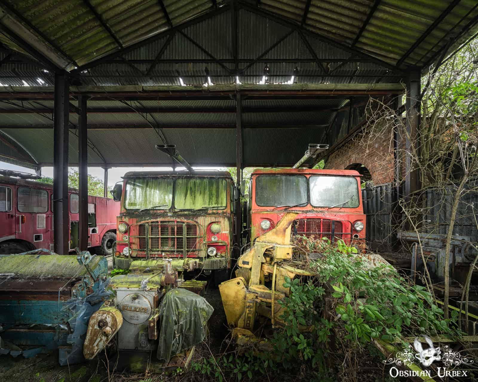 Two abandoned red fire trucks sit in a dilapidated barn, overgrown with moss and foliage. In the foreground, other rusted machinery