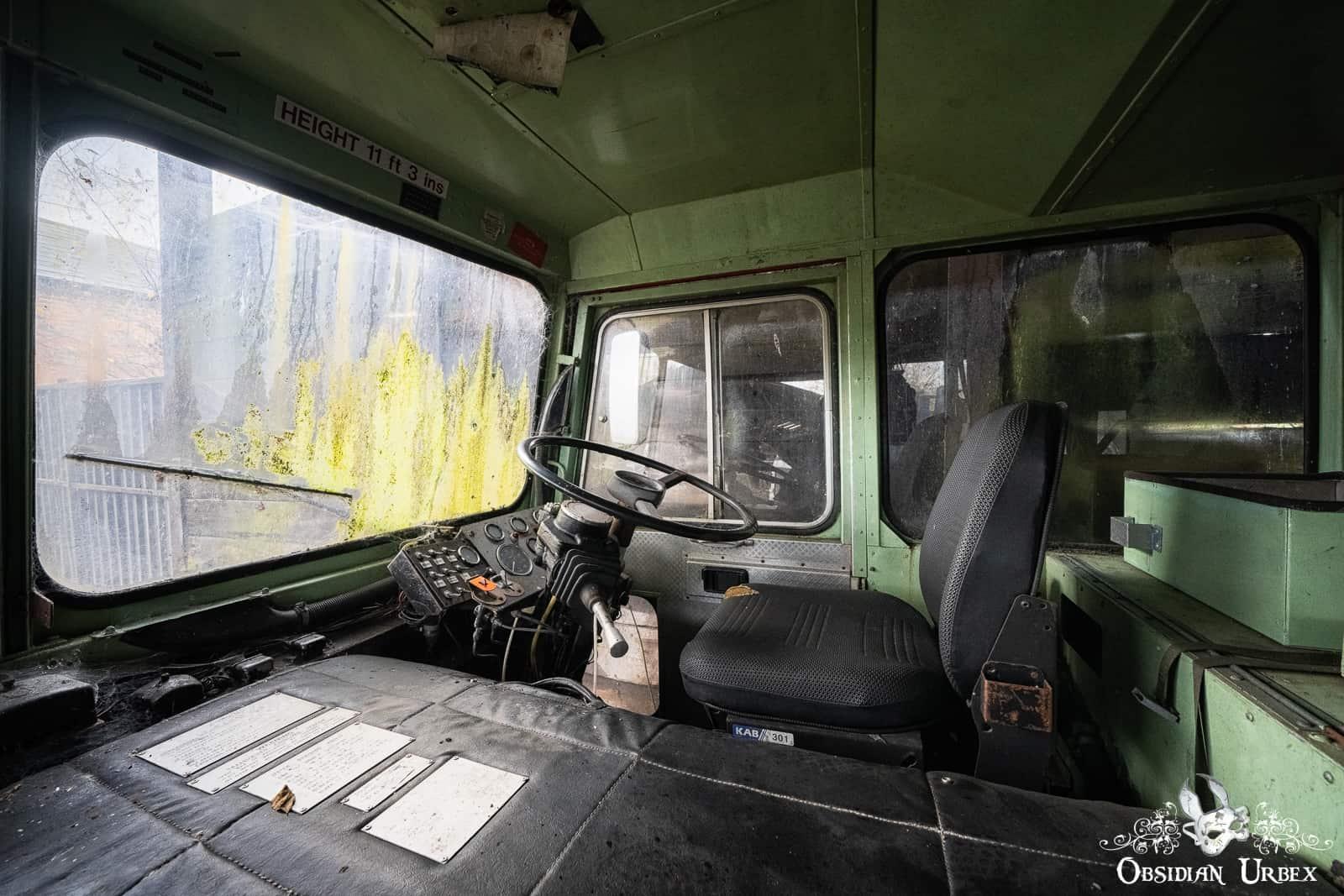 The worn interior of an abandoned green airport fire truck features a driver's seat, steering wheel, and dashboard.