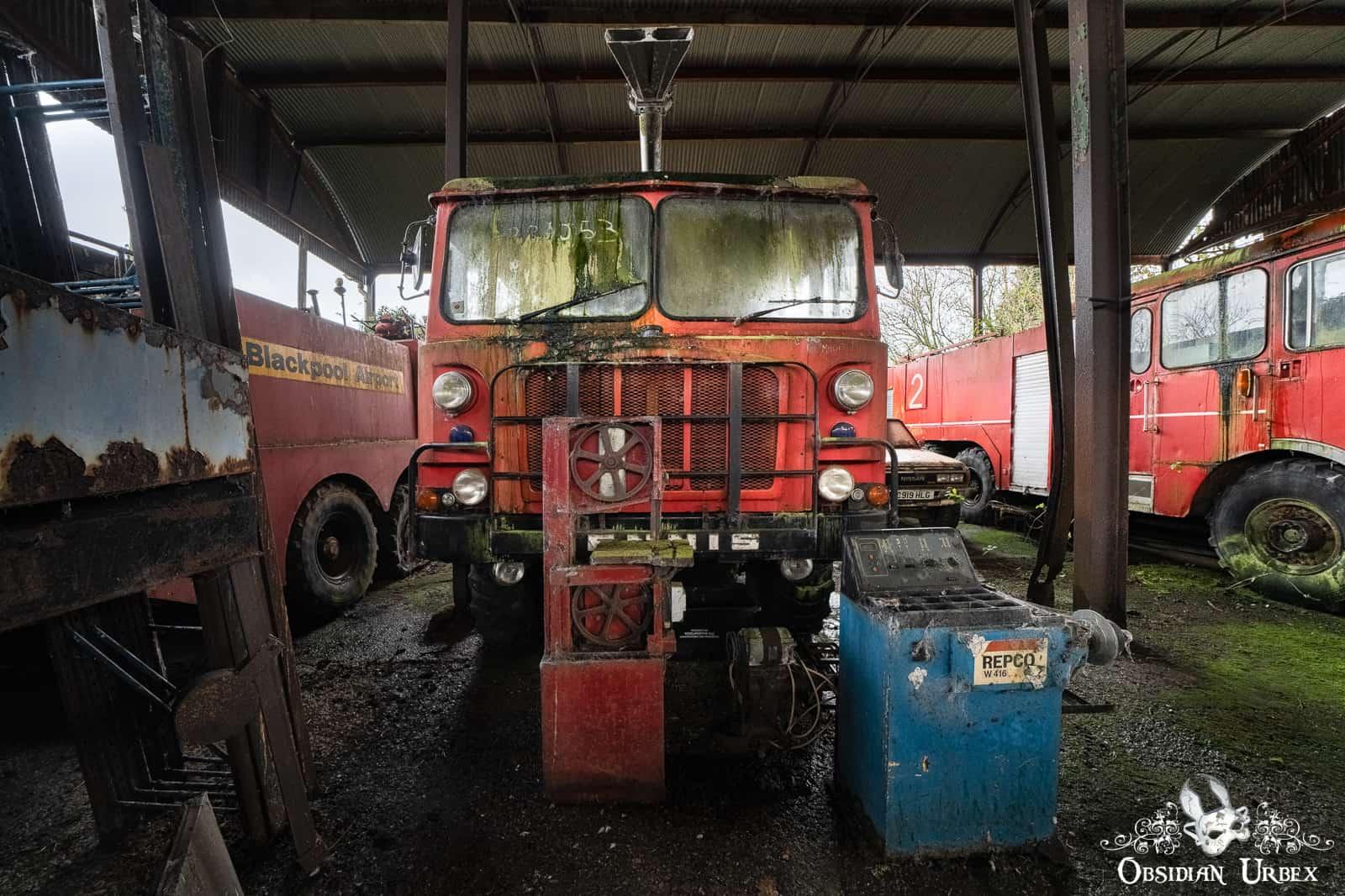 An abandoned, rusty red fire truck, marked "053", sits inside a barn with other old fire trucks.
