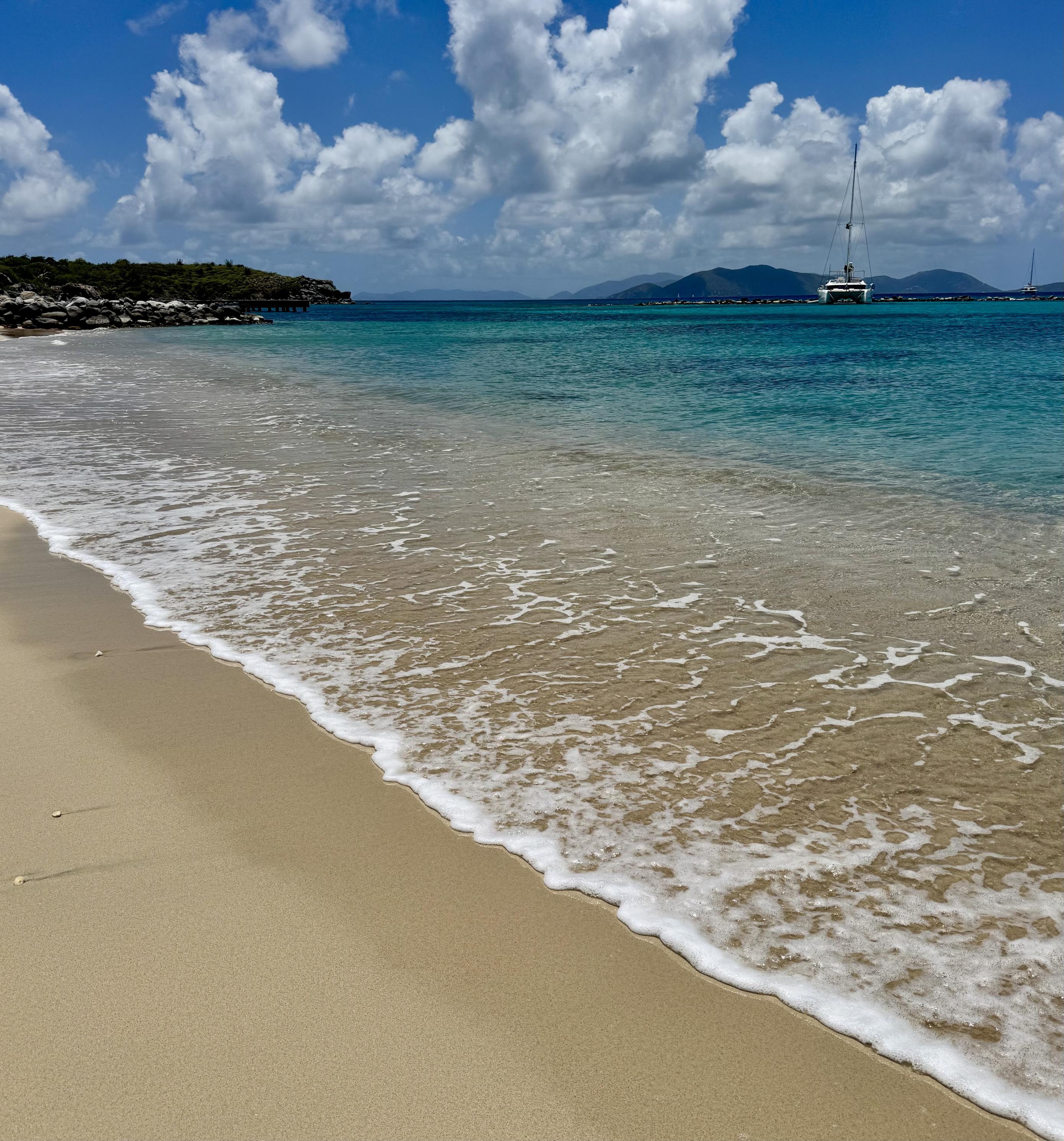 Photo of sandy beach as viewed from land and looking out over the water.  A line of foam from the gentle surf is in the foreground.  Aqua colored water seen offshore, illustrating how clear the water was.  A sailboat and mountainous islands on the horizon.  Partly cloudy skies with patches of blue overhead.

Spanish Town, Virgin Gorda. Looking toward Sir Francis Drake Channel.