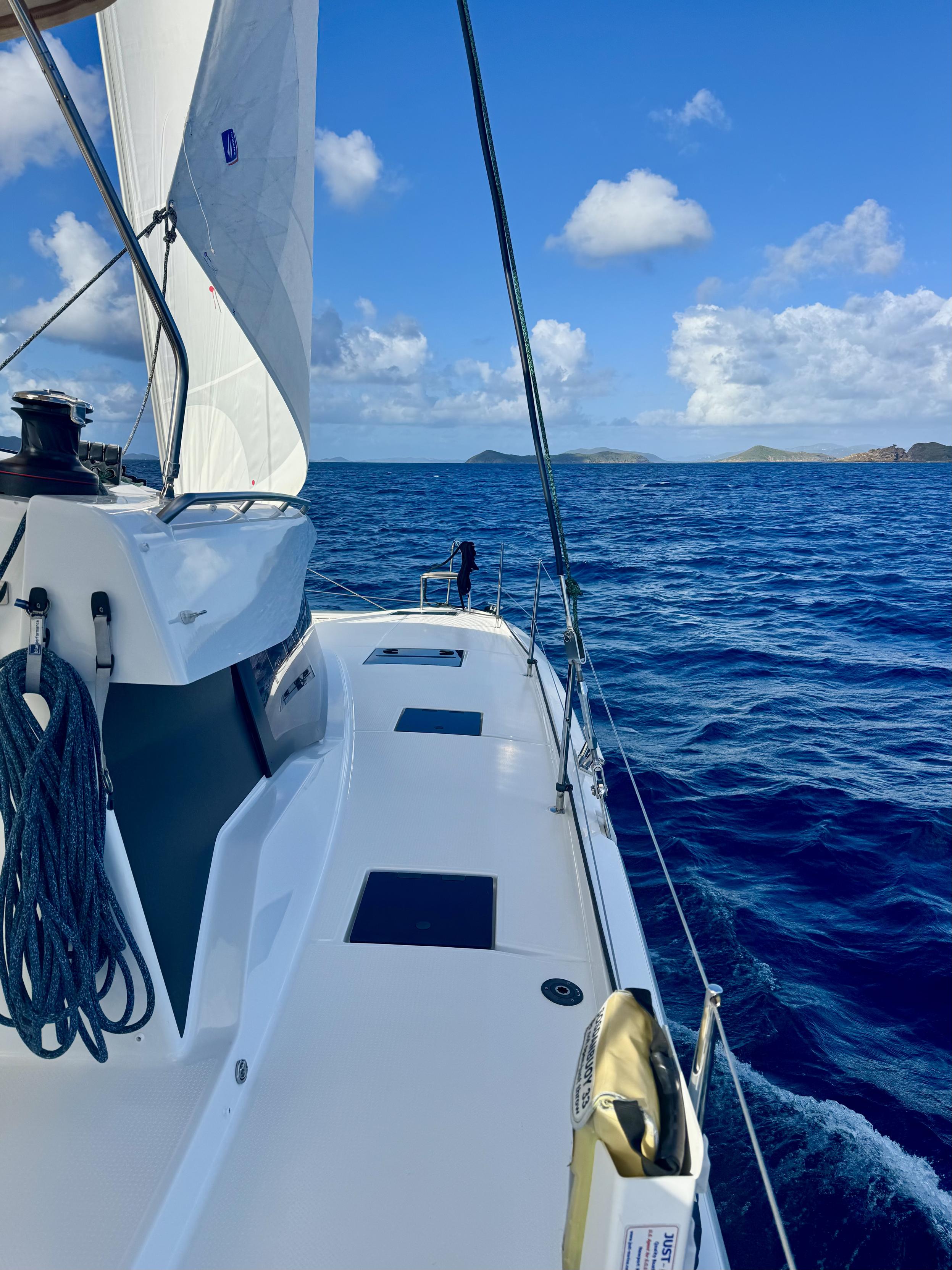 Photo taken from sailboat while sailing.   The forward-most sail (jib) is pulled in tightly.  The boat is white and the view is looking forward as seen from the right (starboard) side of the boat.  Islands can be seen off in the distance.  Partly cloudy sky overhead.

Sir Francis Drake Channel.