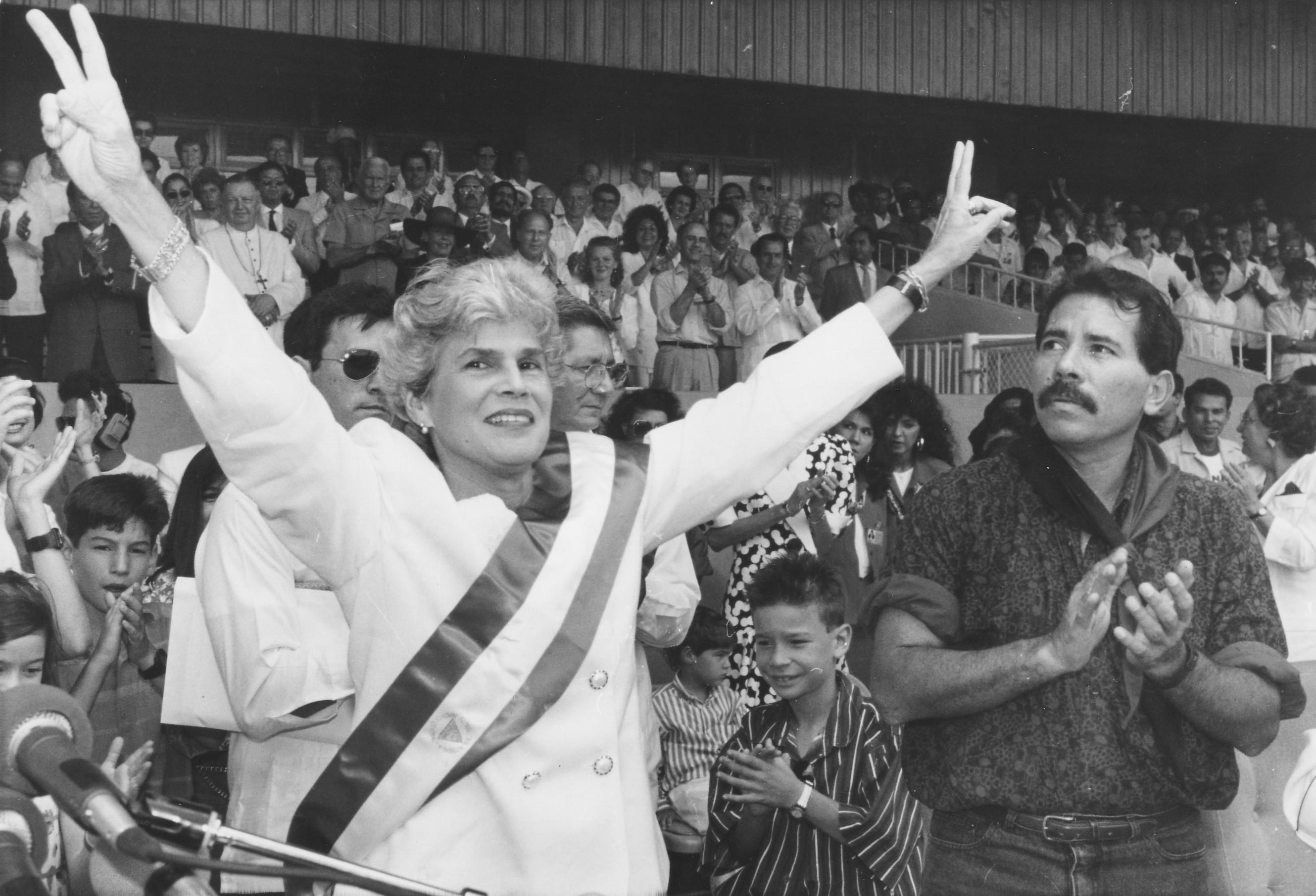 Violeta Barrios de Chamorro wearing the Presidential sash and making V-for-victory signs. Daniel Ortega, a leader of the Sandinistas, is applauding her.