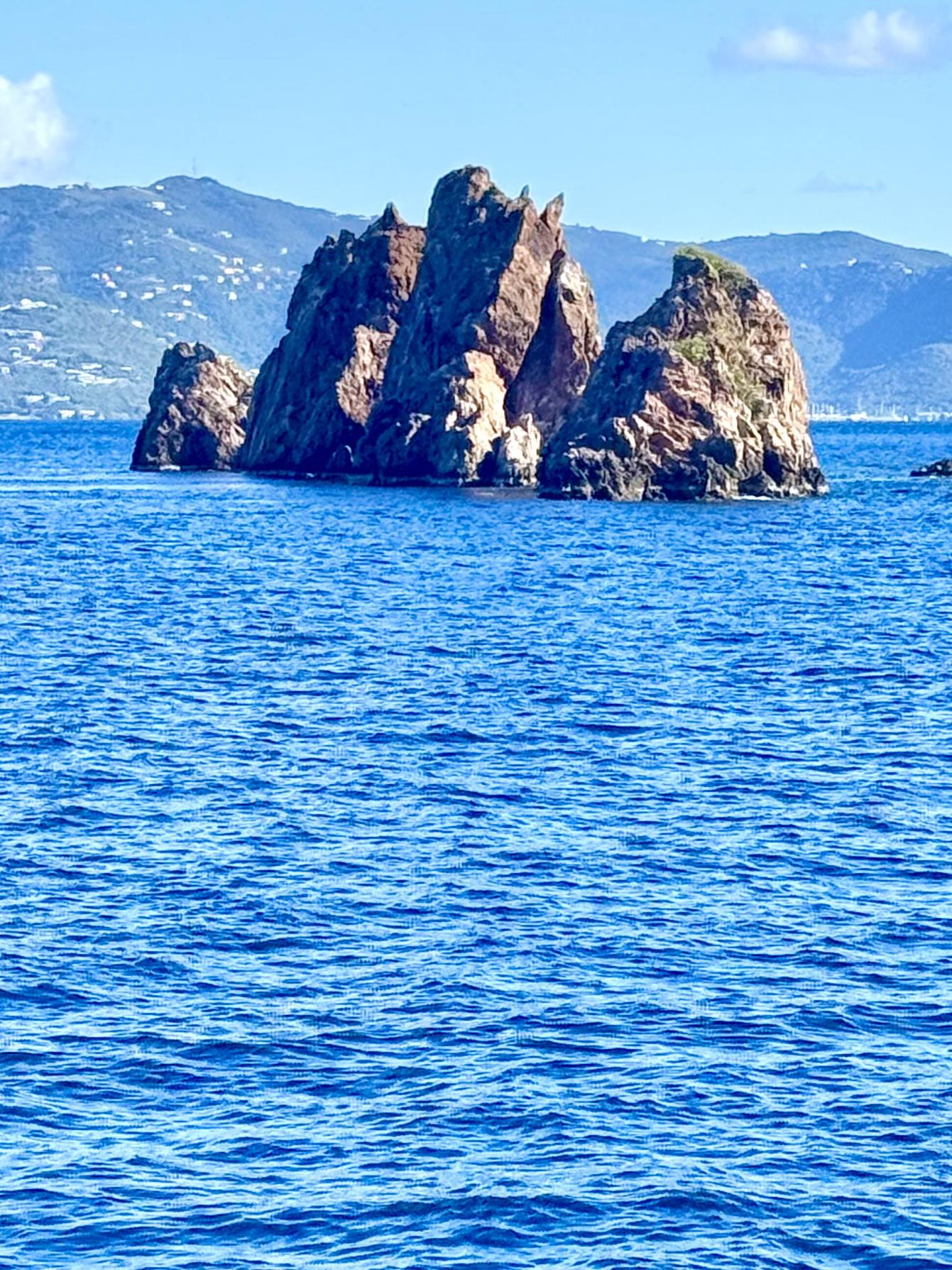 Photo of rock island in ocean. Several closely spaced rock formations jut steeply up out of the water. Larger island in background. Blue sky overhead. 

The Indians, near Norman Island, British Virgin Islands.