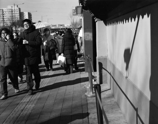 Lucky LUCKY SHD 400 (6x7)

English
A black and white photograph showing a group of people walking on a wooden sidewalk. Several individuals are dressed in winter clothing, including coats and scarves. One person is holding a checkered umbrella. The background features a building and a shadow of a person is visible on the wall.
中文
一张黑白照片，展示了一群人在木板走道上行走。几个人穿着冬季服装，包括外套和围巾。其中一人手持一把格子花纹的雨伞。背景中有一栋带建筑，墙上映出一个人的影子。