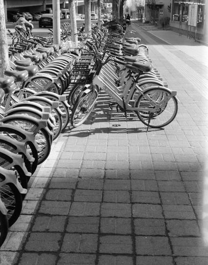 Lucky LUCKY SHD 400 (6x7)

English
A black and white photograph showing a row of bicycles neatly parked in a bike-sharing station. The station is located on a paved sidewalk. The background includes a street lined with trees and buildings, indicating an urban setting.
中文
一张黑白照片，展示了一排整齐停放在共享单车站的自行车。单车站位于铺砌的人行道上。背景中有树木和建筑物，表明这是一个城市环境。