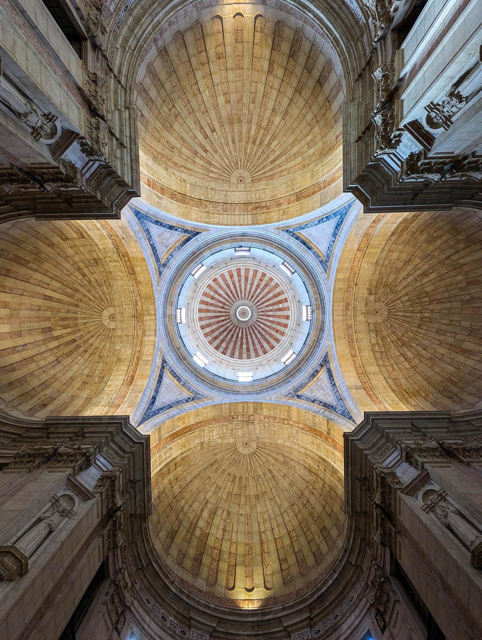 We're inside a huge church-like building at the crossing looking directly up at the dome high above us. The view is perfectly symmetrical. At each corner there are square, stone tower forms reaching up. The space between them has partial domes, warm-toned when compared to the cooler greys of the tower and the overall shape above our heads is a fat Greek Cross (a plus sign) with rounded tips. In the dead centre, directly above our view, there's a full dome, quite small with the distance but well-lit with natural light from windows circling its base. Pale, slightly curved, triangular connecting structures flow out from four points on the exterior of the circular dome to reach the tips of the towers.