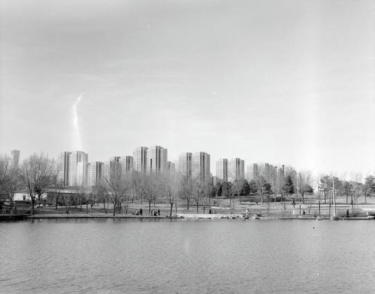 Lucky LUCKY SHD 400 (6x7)

English
A black-and-white photograph of a large urban park with a lake in the foreground. The lake is calm, and the opposite shore is lined with leafless trees. People are scattered around the park, some walking near the water, others sitting on benches. In the background, a skyline of tall apartment buildings is visible, indicating the proximity of the city.
中文
一张黑白照片，展现了一个大型城市公园，前景是一个湖泊。湖面平静，对岸种满了光秃秃的树木。公园里散布着一些人，有的在水边散步，有的坐在长椅上。背景中，高层公寓楼的天际线清晰可见，表明公园靠近市区。