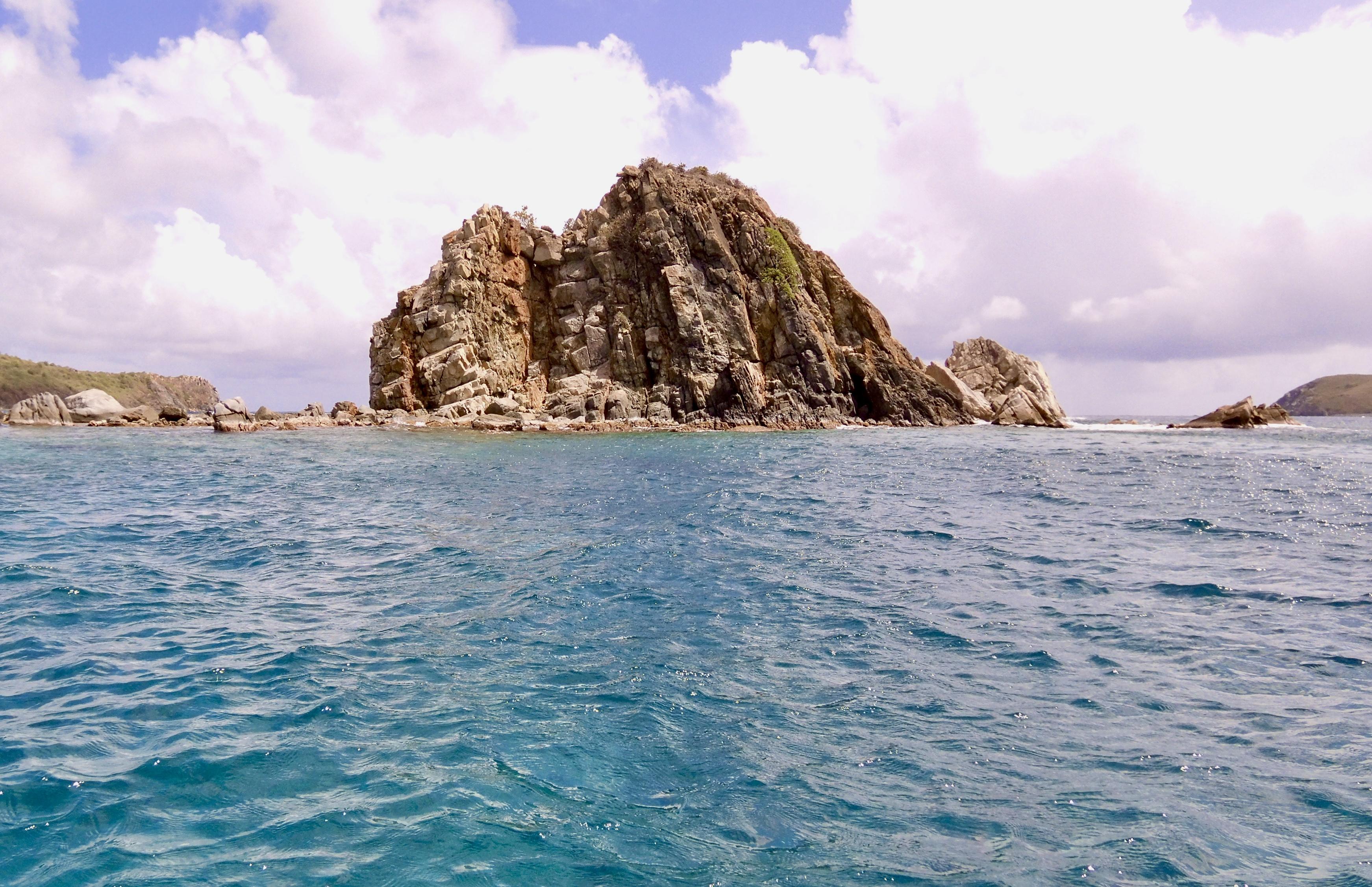 Photo of rock island as viewed from the dinghy.  I used the dinghy from the sailboat to get close enough to the island to scuba dive.  The island is solid rock and is seen jutting steeply up out of the water.  Other land masses are seen behind it.  Mostly cloudy skies with small patches of blue overhead.

Cistern Point, Cooper Island, British Virgin Islands.