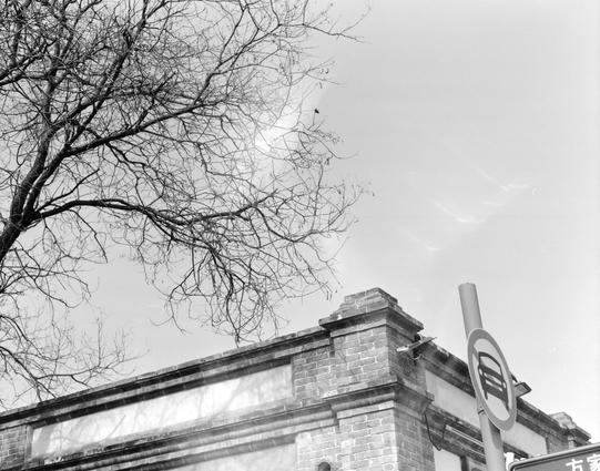 Kentmere PAN 200 (6x7)

English
A black-and-white photograph showing the upper corner of a brick building with a traffic sign on the right side. The building has a flat roof and is partially covered by bare tree branches extending from the left side of the frame. The sky is clear and bright, providing a stark contrast to the building and branches.
中文
一张黑白照片，展示了一栋砖砌建筑的上角，右侧有一个交通标志。建筑物屋顶平整，左侧伸出的光秃树枝部分遮挡了建筑。天空清朗明亮，与建筑和树枝形成鲜明对比。