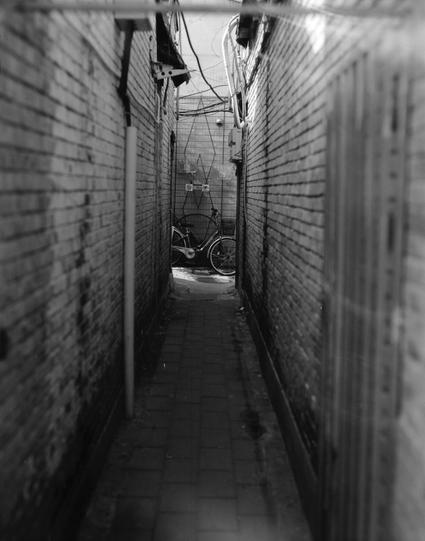 Kentmere PAN 200 (6x7)

English
A black-and-white photograph of a narrow alleyway between two brick walls. The alley is paved with bricks and leads to a dead end where a bicycle is parked against the wall. Overhead, electrical wires and cables are visible, stretching across the alley. The walls are tall and narrow, creating a sense of depth and confinement.
中文
一张黑白照片，展示了两面砖墙之间的狭窄小巷。小巷铺着砖，尽头处有一辆自行车靠墙停放。头顶上方可见电线和电缆横跨小巷。墙壁高而窄，营造出深度和局促感。