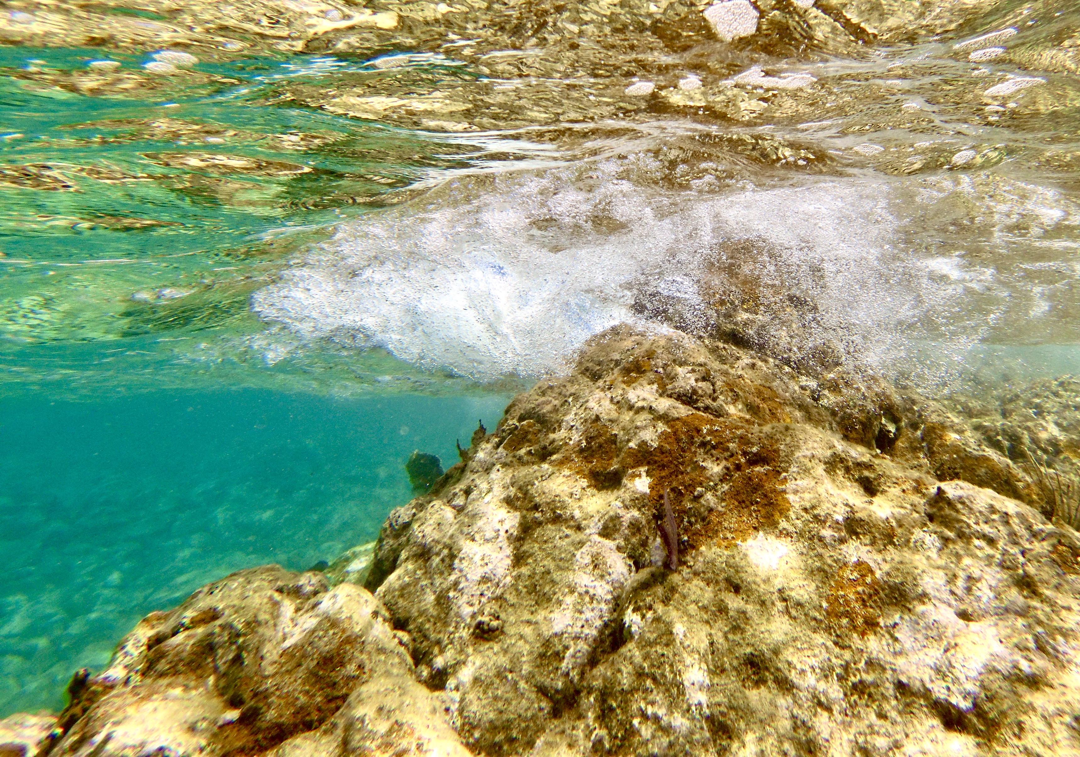 Photo taken underwater looking up at surface of water.  The water is seen crashing over a large rock formation.  A dense cloud of bubbles surrounds the peak of the rock formation.  The water surface is reflective in the foreground.  The water is greenish in the background

I took this photo at the point of land at one of the ends of Kelly's Cove, Norman Island, British Virgin Islands.  The point of land is exposed to the open ocean and is conducive to wave action with an onshore wind.