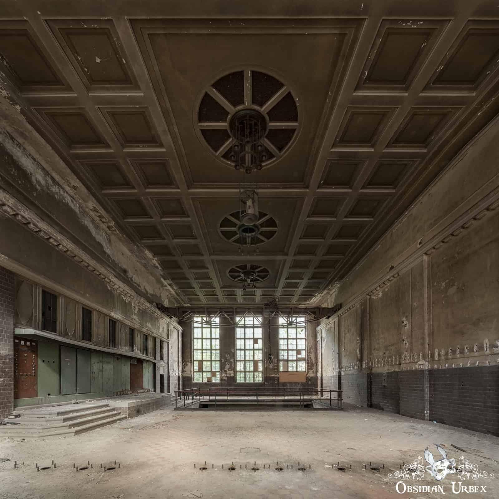 A grand, abandoned industrial hall features a coffered ceiling with circular vents and tall windows at the far end. The space shows decay, with dirt on the floor and peeling paint on the walls.