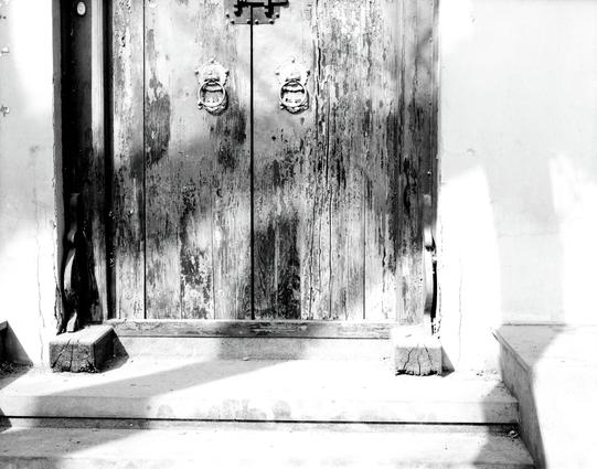 Kentmere PAN 200 (6x7)

##English
A black-and-white close-up of an old, weathered wooden door. The door features two circular metal door knockers and a vertical metal handle. The wood shows signs of age, with visible cracks and a worn surface. The door is set within a stone frame, and sunlight casts sharp shadows on the ground in front of it.
##中文
一张黑白特写照片，展示了一扇古老、风化的木门。木门上有两个圆形的金属门环和一个垂直的金属把手。木头显示出岁月的痕迹，表面有可见的裂缝和磨损。木门嵌在一个石头门框内，阳光在门前的地面上投射出清晰的影子。
