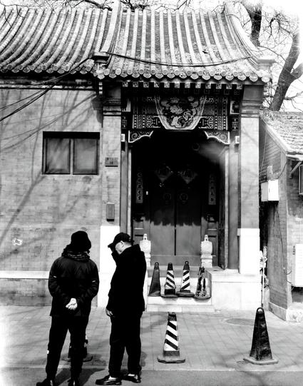 Kentmere PAN 200 (6x7)

##English
A black-and-white photograph of the entrance to a traditional East Asian building. The entrance is framed by a richly decorated archway with intricate carvings and a tiled roof. Two individuals stand in front of the entrance, engaged in conversation. Traffic cones are placed near the entrance, possibly indicating restricted access. The scene captures a blend of historical architecture and modern elements.
##中文
一张黑白照片，展示了传统东亚建筑的入口。入口由一个装饰精美的拱门框起，拱门上有复杂的雕刻和瓦屋顶。两个…