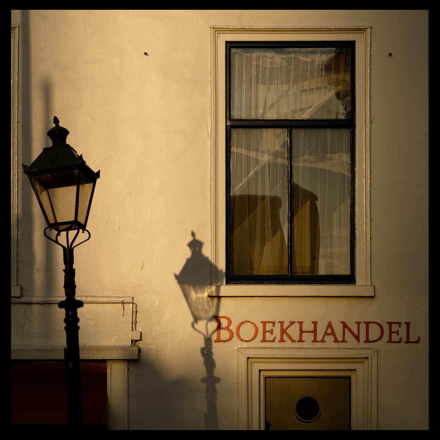 A colour photograph of an out of kilter, vintage lamppost casting a shadow on the wall. In the top right-hand corner, a window can be seen casring reflections. The word Boekhandel (Bookshop) is written on the wall in red letters. Everything is bathed in the warm morning light of the rising sun.
 
Camera: Nikon Z6ii
Lens: Nikkor AF-S 85mm f/1.4G