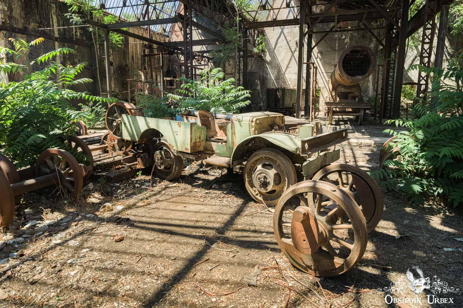 An abandoned industrial interior features a rusty light-green 4x4, scavenged for parts, amidst detached wheels and lush green foliage. The setting is an old train depo