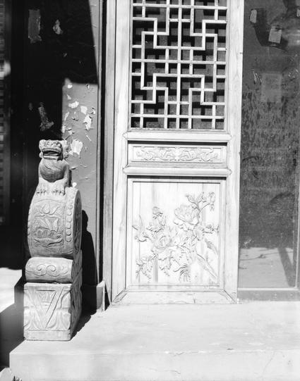 Kentmere PAN 200 (6x7)

##English
Black-and-white close-up of a traditional Chinese door with intricate geometric lattice windows. To the left of the door stands a carved stone lion statue on a pedestal, featuring detailed patterns. The door panel below the lattice displays a carved floral design.
##中文
一张黑白特写照片，展示了一扇传统中式门，门上有精美的几何格子窗。门的左侧有一尊雕刻精细的石狮子像，立在一个带有细节图案的基座上。格子窗下方的门板上展示了一个雕刻的花卉图案。