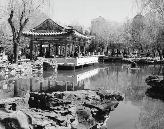 Kentmere PAN 200 (6x7)

##English
Black-and-white photograph of a traditional Chinese pavilion with ornate, upturned eaves, situated beside a calm pond. The pavilion is elevated on a stone platform, connected by a bridge. Rocky formations and bare trees surround the water, with a few people visible near the structure. In the background, modern buildings are faintly visible through the trees.
##中文
一张黑白照片，展示了一座传统中式凉亭，亭子屋顶翘角装饰精美，位于一个宁静的池塘旁。凉亭建在石头平台上，通过一座桥与对岸相连。周围是岩石地貌和光秃的树木，亭子附近有几个人。远处透过树木可以隐约看到现代…