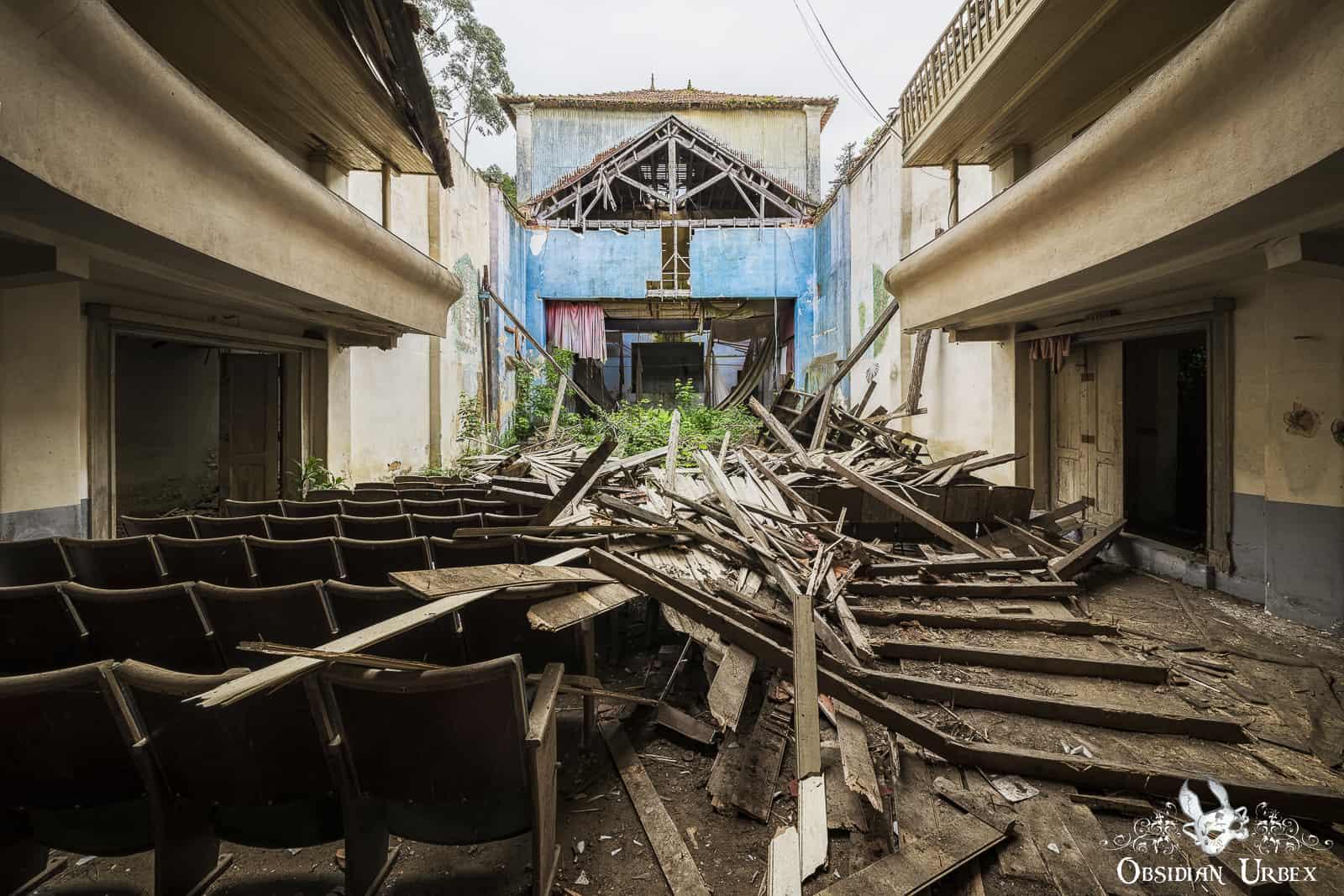 An abandoned theater shows rows of seats, with a collapsed roof and debris scattered across the floor leading to an overgrown stage with blue walls.
