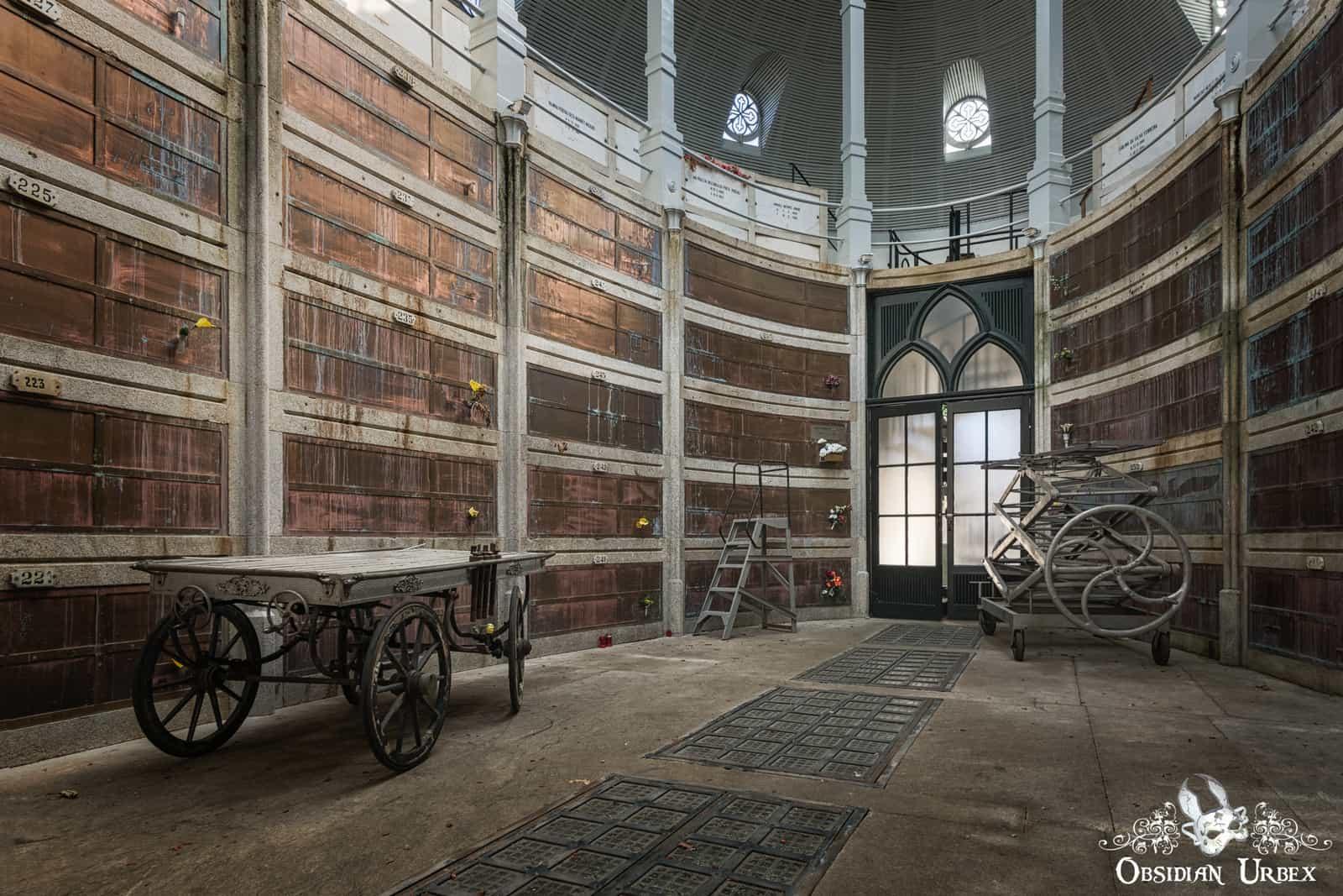 An abandoned circular crypt has walls lined with bronze burial niches, some numbered. Two antique wheeled carts and a ladder sit on the concrete floor near an arched door.