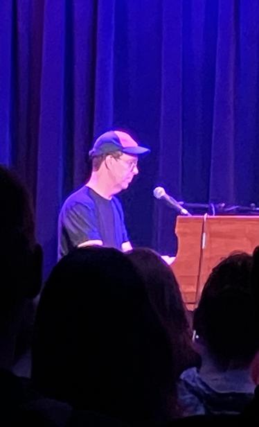 Ben Folds is playing a piano on stage, wearing a cap and glasses, with a microphone in front of him. Behind him, dark curtains are illuminated by purple lighting. The blurred silhouettes of audience members are visible in the foreground.