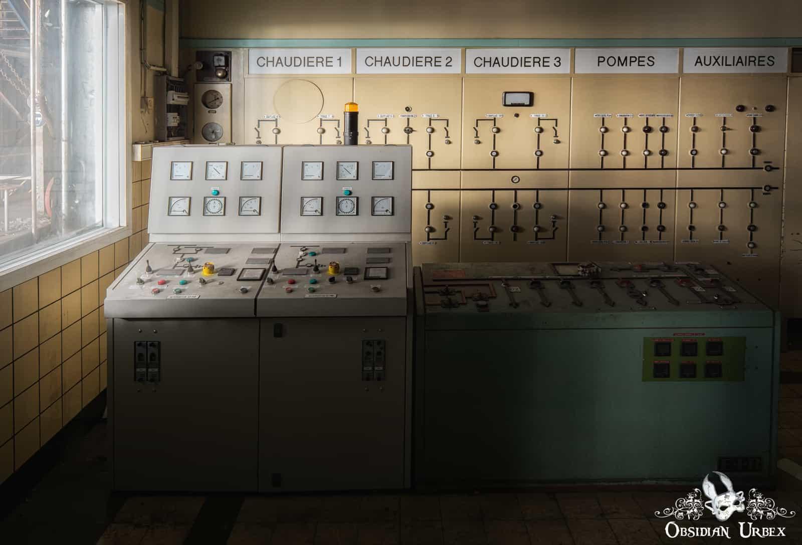 An abandoned industrial control room features two large consoles and a wall panel labeled CHAUDIERE 1, CHAUDIERE 2, CHAUDIERE 3, POMPES, AUXILIAIRES.