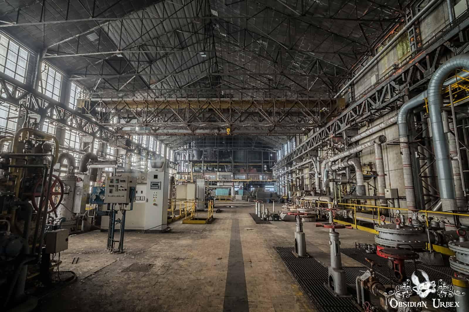 A vast, derelict turbine hall inside an abandoned power plant, filled with industrial machinery, pipes, and control panels.