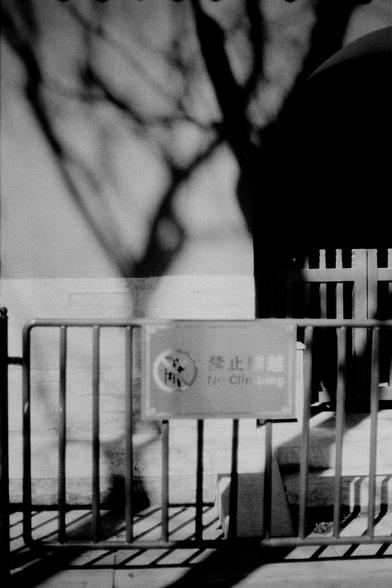 ERA 100 (FF)

English
Black-and-white photograph of a metal barricade with a sign attached. The sign reads "禁止翻越" (No Climbing) in Chinese characters, with the English translation "No Climbing" beneath. The barricade is positioned on a paved surface, with tree shadows cast across the scene.
中文
黑白照片，拍摄的是一个金属栅栏，上面附有标志。标志上用中文写着“禁止翻越”，下方有英文翻译“No Climbing”。栅栏位于铺设的地面上，树木的影子投射在场景中。