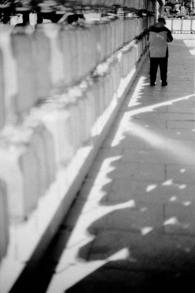 ERA 100 (FF)

English
Black-and-white photograph of a person standing on a paved walkway beside a set of stacked lanterns. The person is facing away from the camera, wearing a light-colored jacket and dark pants. The lanterns cast a shadow on the ground, and the foreground is slightly blurred, emphasizing the person and the lanterns next to the person.
中文
黑白照片，拍摄的是一个站在铺设人行道上的人，旁边是灯笼和架子。此人背对着相机，穿着浅色夹克和深色裤子。灯笼在地面上投下影子，前景略微模糊，突出了人物和其身旁的灯笼。