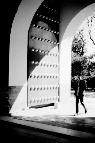 ERA 100 (FF)

English
Black-and-white photograph of a person walking past a large, arched gateway. The gateway is made of light-colored stone and features a massive, dark, riveted metal door with rows of decorative rivets. The person, dressed in a jacket and pants, is seen from the side, holding a phone or device. The scene is set in a park or garden, with trees visible in the background.
中文
黑白照片，画面中一人正从一个大型拱形门前走过。拱门由浅色石头建造，门上是一扇巨大的深色铆钉金属门，排列着装饰性铆钉。人物身穿夹克和裤子，从侧面可见，手中拿着手机或设备。背景是公园或花园的景象，可看到树木。