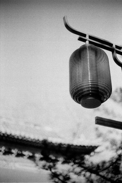 ERA 100 (FF)

English
Black-and-white photograph of a traditional, cylindrical lantern hanging from a curved metal arm. The lantern has a ribbed texture and a dark, circular base. The background features a blurred sky and the silhouettes of tree branches and a traditional Chinese roof top with eaves, creating a serene, atmospheric scene.
中文
黑白照片，画面中一个传统的圆柱形灯笼悬挂在一个弯曲的金属臂上。灯笼表面有肋状纹理，底部为深色圆形。背景是模糊的天空和树枝以及传统中国房顶屋檐的剪影，营造出一种宁静的氛围。