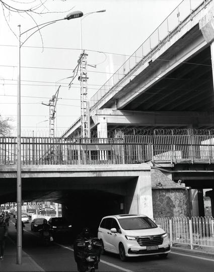 Kentmere PAN 400 (6x7)

##English
Black and white photograph of an urban scene featuring a concrete overpass with a metal railing on top. Below the overpass, a white Honda SUV with the license plate "京HD819" is driving on the road. A motorcyclist is visible in the foreground. The overpass is supported by large concrete pillars, and utility poles with streetlights and electrical wires are present. In the background, there is a fence with barbed wire and a partially visible elevated structure. Ev…
