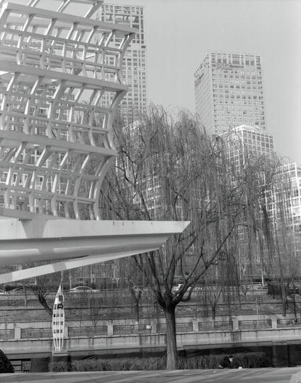 Kentmere PAN 400 (6x7)

Black and white photograph of a modern urban area. The foreground features a large, intricate white metal structure with geometric patterns. Behind it, tall buildings with numerous windows are visible. Bare trees with thin branches are scattered throughout the scene, adding a natural element to the otherwise architectural focus.
##中文
黑白现代城市景观照片。前景是一个大型、复杂的白色金属结构，具有几何图案。其后是多个高层建筑，建筑上有众多窗户。画面中还零星分布着光秃秃的树木，细细的树枝为以建筑为主的景观增添了一丝自然元素。