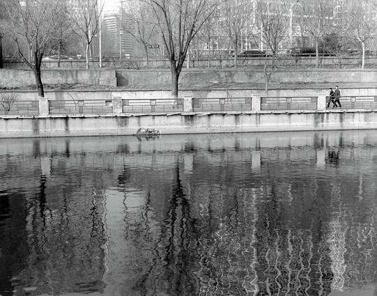 Kentmere PAN 400 (6x7)

English
Black and white photograph of a calm river with ripples on the water surface. On the opposite bank, there is a concrete embankment with a railing and a row of leafless trees. Two people are walking along the embankment. In the background, buildings and a bridge are visible, reflecting slightly in the water.
中文
黑白照片展示了一条平静的河流，水面有涟漪。对岸是一个带有栏杆的混凝土堤岸，以及一排光秃秃的树木。两个人正在堤岸上行走。背景中可以看到建筑物和一座桥，在水中有轻微的倒影。