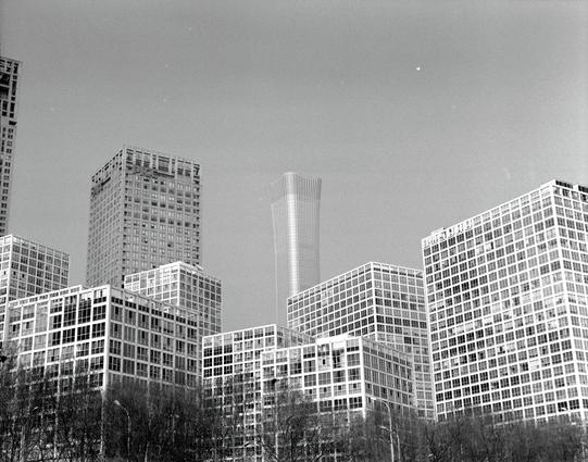 Kentmere PAN 400 (6x7)

English
Black and white photograph of a city skyline featuring multiple high-rise buildings. The tallest structure is a modern, uniquely shaped skyscraper with a twisted design, standing prominently in the center. Surrounding it are several rectangular buildings with numerous windows, arranged in a semi-circle. The foreground includes bare trees, suggesting a winter or early spring setting.
中文
黑白照片展示了一个城市的天际线，包含多栋高层建筑。最高的建筑是一座现代化的摩天大楼，具有独特的扭曲设计，位于画面中央。周围是多栋带有众多窗户的矩形建筑，呈半…