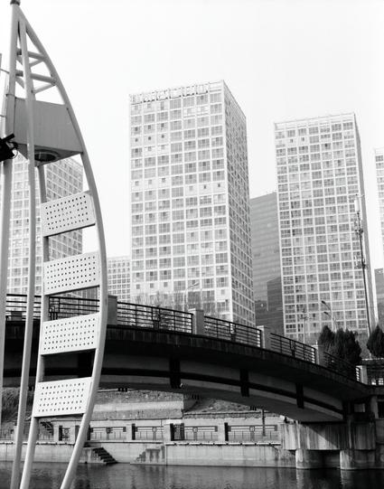 Kentmere PAN 400 (6x7)

English
Black and white photograph of a modern pedestrian bridge with a curved design. The bridge has railings on both sides and connects to a tall, white structure. In the background, there are several high-rise buildings with numerous windows, reflecting on the water below.
中文
黑白照片展示了一座现代化的行人桥，设计为弯曲形状。桥的两侧都有栏杆，左侧连接到一个高大的白色结构。背景中有多栋高层建筑，窗户众多，倒影在下方的水面上。