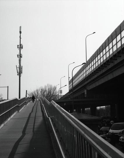 Kentmere PAN 400 (6x7)

English
Black and white photograph of a long, elevated walkway with railings on both sides. A single person is walking away from the camera. On the right, there is a tall overpass with streetlights and vehicles visible underneath. On the left, there is a tall pole with multiple devices attached, likely antennas or cameras. Bare trees are visible in the background.
中文
黑白照片展示了一条长长的高架人行道，两侧都有栏杆。一个人正背对着相机走远。右侧是一座高大的立交桥，可以看到路灯和下方的车辆。左侧有一根高杆，上面安装了多个设备，可能是天线或摄像头。背景中可以看到光秃秃的树木。