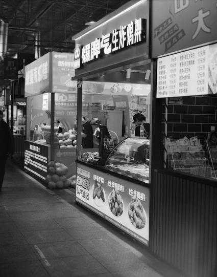 Kentmere PAN 400 (6x7)

##English
A black-and-white photograph of a modern food stall in a market. The stall features illuminated signage with Chinese characters. The display case is filled with various food items, and a menu with prices is visible on the right side. The stall is clean, well-organized, and inviting, with a few customers visible in the background.
##中文
一张黑白照片，展示了市场里的一家海鲜摊位。摊位上方有中文发光招牌，展示柜中摆满了各种食物。右侧可见一张带价格的菜单。摊位干净、整洁且吸引人，背景中还能看到几位顾客。