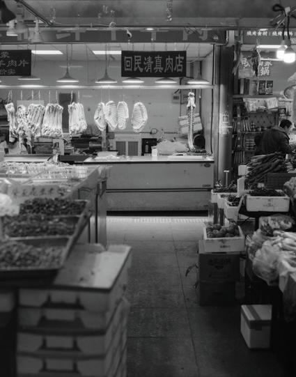 Kentmere PAN 400 (6x7)

##English
A black-and-white interior shot of a traditional meat shop. The shop's signboard reads "回民清真肉店" (Halal Meat Shop). Several pieces of meat, likely lamb or beef ribs, hang from hooks in the foreground. The shop is well-lit, with a glass display case and various items visible inside. The scene conveys a busy, authentic market atmosphere.
##中文
一张黑白室内照片，展示了一家传统的肉店。店铺的招牌上写着“回民清真肉店”。前景中有几块肉，可能是羊排或牛排，挂在钩子上。店内灯光明亮，有一个玻璃展示柜，里面还能看到各种物品。场景传达了忙碌、地道的市场氛围。