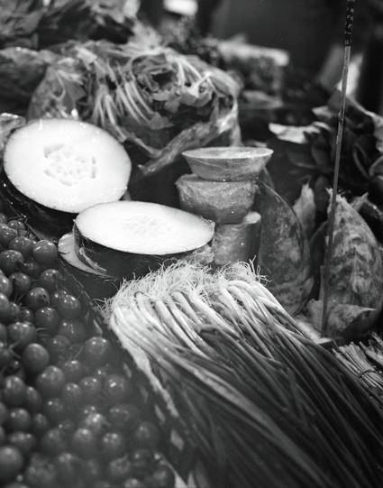 Kentmere PAN 400 (6x7)

##English
A black-and-white close-up of a market stall displaying various food items. In the foreground, there are large, halved melons. To the right, a stack of cut pumpkins is visible. In the background, bundles of dried herbs or plants hang, and a pile of round, dark-colored fruits or vegetables is present. The scene is bustling with textures and traditional market elements.
##中文
一张黑白近景照片，展示了市场摊位上的各种食品。前景是两个被切开的冬瓜。右侧是一叠小小的切开的南瓜。后方悬挂着束干燥的草药或植物，还有一堆深色的圆形水果或蔬菜。场景充满了质感和传统…
