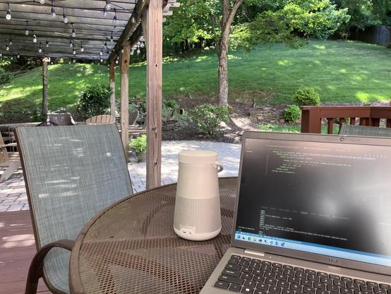 A outdoor workspace featuring a laptop displaying code on a table, alongside a white speaker. There is a chair in the foreground, and a well-maintained grassy area with trees and decorative lights in the background.