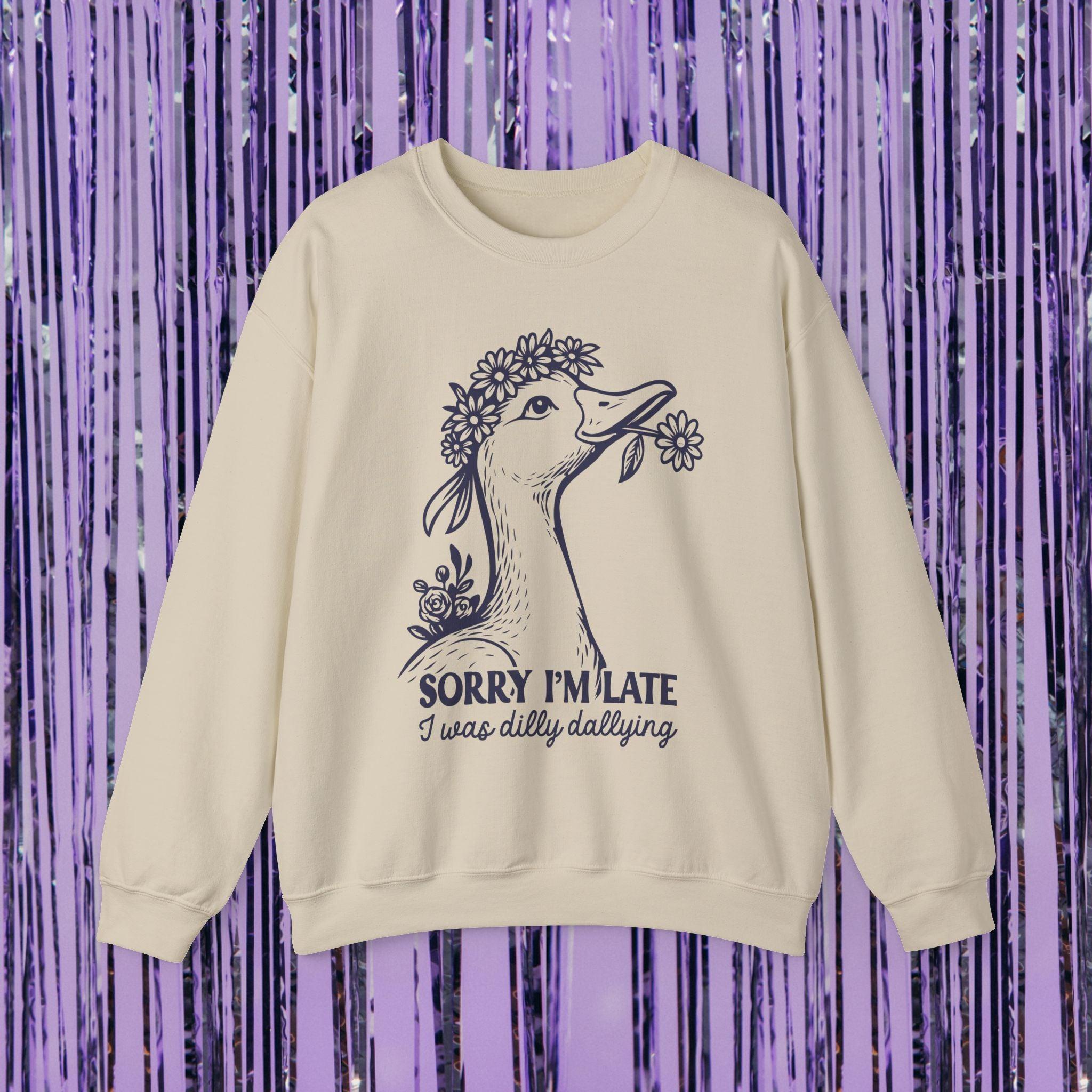 a cream-colored crewneck sweatshirt is displayed against a purple tinsel backdrop. The sweatshirt features a central graphic of a duck wearing a flower crown and holding a daisy in its beak. Below the illustration, the text reads, "SORRY I'M LATE I was dilly dallying."