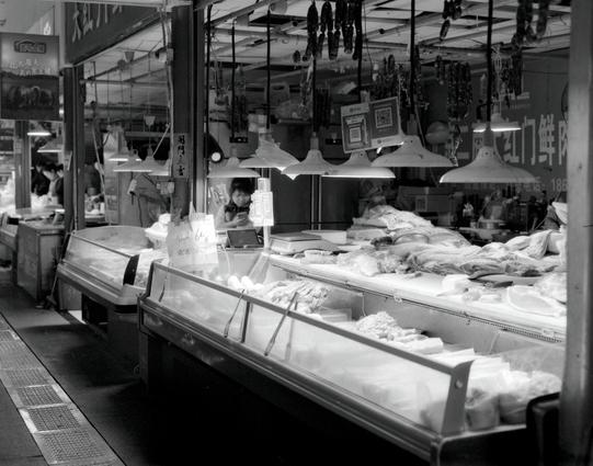 Kentmere PAN 400 (6x7)

English
A black-and-white photograph of a butcher’s stall. The stall has a glass display case filled with various types of meat and other products. Hanging lamps illuminate the stall, and there are signs with text, including a QR code. The background shows more market activity and hanging dried goods.
中文
一张黑白照片，展示了一个肉摊。摊位有一个玻璃展示柜，里面摆放着各种肉类和其他产品。悬挂的灯照亮了摊位，还有带文字的标志，包括一个二维码。背景显示了更多市场活动和悬挂的干货。