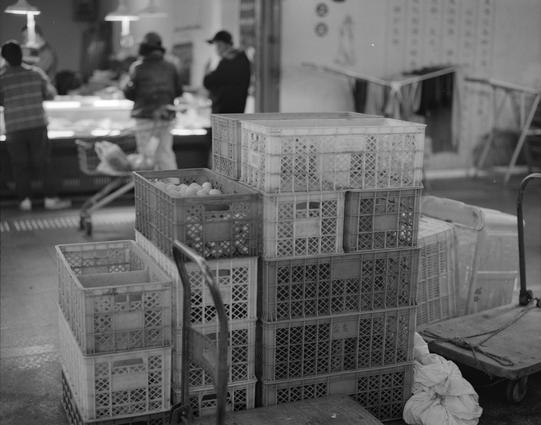 Kentmere PAN 400 (6x7)

English
A black-and-white photograph of a bustling indoor market. In the foreground, there are several stacked plastic crates, some containing eggs. The background shows blurred figures of people, likely vendors and customers, engaged in various activities. The market appears well-lit with hanging lamps visible in the ceiling area.
中文
一张黑白照片，展示了一个繁忙的室内市场。前景是几个叠放的塑料箱，其中一些箱子里装着鸡蛋。背景中可以看到模糊的人影，可能是商贩和顾客，他们正在进行各种活动。市场内光线充足，天花板上悬挂着多盏灯。