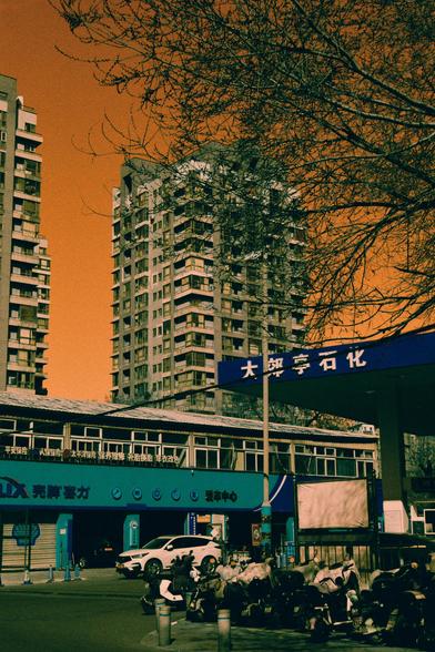 Harman Switch Azure (FF)

nglish
A cityscape with an orange-tinted sky. In the foreground, there is a blue and white building with Chinese characters on its facade. Below the sign, there are smaller Chinese characters listing various services. Several motorbikes are parked in front of the building. A white car is also visible. Tall residential buildings rise in the background, with bare tree branches in the foreground.
中文
一幅城市景观，天空呈现橙色色调。前景是一栋蓝白相间的建筑，其立面有中文字样。招牌下方还有更小的中文字样，列出了各种服务。建筑前停放着多辆摩托车，还…