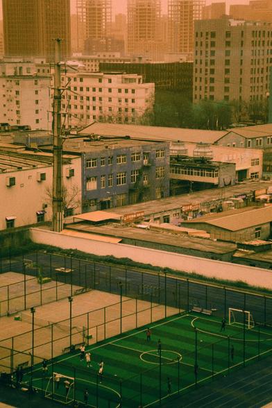 Harman Switch Azure (FF)

English
An aerial view of an urban area with a mix of old and new buildings. In the foreground, there is a green artificial turf sports field with a few people playing. The field is enclosed by a fence. Behind it, there are low-rise buildings with flat roofs, some of which appear worn. Tall modern skyscrapers loom in the distant background under a hazy orange sky.
中文
俯瞰城市地区，新旧建筑混杂。前景是一个绿色人造草皮运动场，有几个人在活动，场地被围栏围起。后方是一些低矮的平顶建筑，部分看起来有些破旧。远处高大的现代摩天大楼在朦胧的橙色天空下若隐若现。