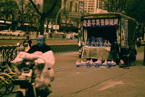 Harman Switch Azure (FF)

##English
A street scene with a truck parked on the right, covered with a tarp and displaying a sign in Chinese characters that reads "吉县苹果香酥脆甜" (Jixian apples, fragrant, crisp, and sweet). Bags of apples are stacked in front of the truck. A person on a bicycle loaded with large white sacks is in the foreground. The background shows a busy urban street with trees, cars, and pedestrians.
##中文
街道场景，右侧停着一辆覆盖着防水布的卡车，车上挂着中文招牌，写着“吉县苹果香酥脆甜”。卡车前方堆放着多袋苹果。前景中有一人骑着自行车，车上装满大白袋。背景是…